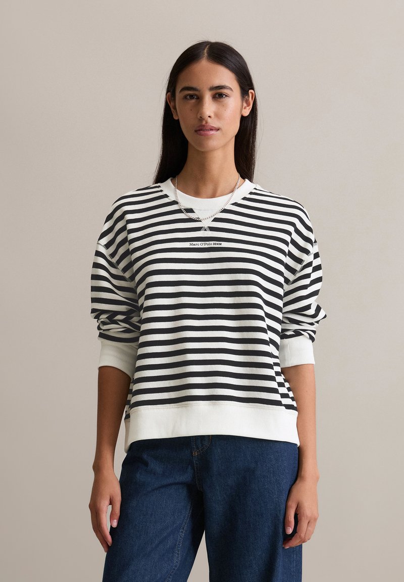 Frau mit schwarzem und weißem gestreiften Langarmshirt mit dem Text "Marc O'Polo Denim" und blauen Jeans, vor einem einfarbigen Hintergrund stehend.
