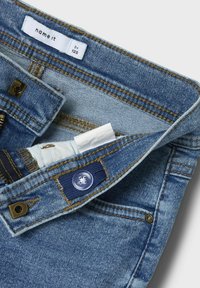 Jeans en denim bleu clair avec une texture décolorée, fermeture éclair argentée, coutures marron et un accent de taille bleu marine. L'étiquette indique "name it by 128."