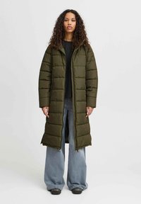 Manteau matelassé long vert olive avec capuche, présentant un motif matelassé, une fermeture éclair à l'avant et des poches latérales. Porté sur un haut noir et un jean coupe large.