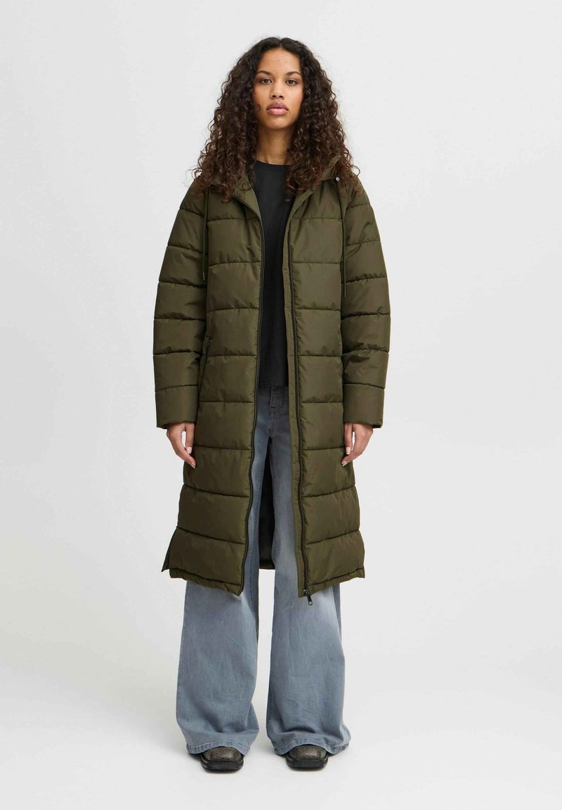 Manteau matelassé long vert olive avec capuche, présentant un motif matelassé, une fermeture éclair à l'avant et des poches latérales. Porté sur un haut noir et un jean coupe large.