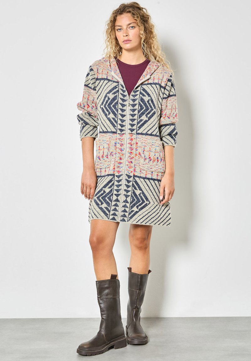 Sweter z kapturem w kolorze kremowym z niebieskimi geometrycznymi wzorami i różowymi akcentami. Posiada długie rękawy, zamek z przodu i sięga do połowy uda. Brązowe botki do kolan.