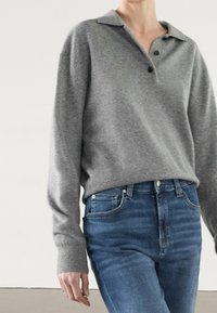 Grå stickad tröja med krage och tre knappar, matchad med hög midja blå jeans. Mjuk textur synlig i tyget.