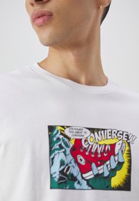 Camiseta blanca con un gráfico colorido de estilo cómic que muestra una mano sosteniendo una zapatilla roja y un texto en negrita que dice "CONVERSE!!".