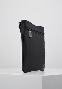 Černá hladká kožená taška se zipovým uzávěrem a nenápadným logem, s elegantním, minimalistickým designem a plochým obdélníkovým tvarem.