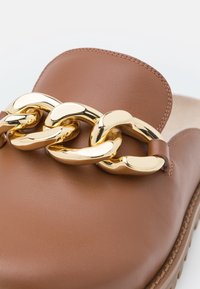 Chaussure en cuir marron à enfiler avec un grand détail en chaîne dorée brillante sur le dessus et des coutures visibles le long des bords.