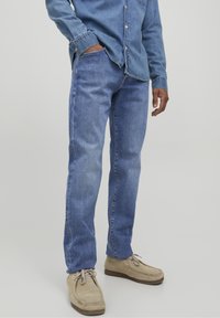 Jeans de mezclilla azul claro con un diseño de pierna recta, que presentan un sutil desgaste y bolsillos laterales, combinados con zapatos beige y una camisa de mezclilla.
