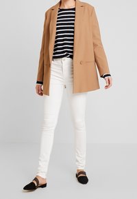 Blazer beige, top a righe verticali blu navy e bianche a maniche lunghe, jeans skinny bianchi e scarpe basse nere con una fascia sul ponte del piede.