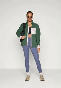 Veste zippée en polaire de couleur vert foncé avec des accents rose clair ; soutien-gorge de sport noir ; leggings bleus ; porte un sac à dos noir ; porte des lunettes de soleil.