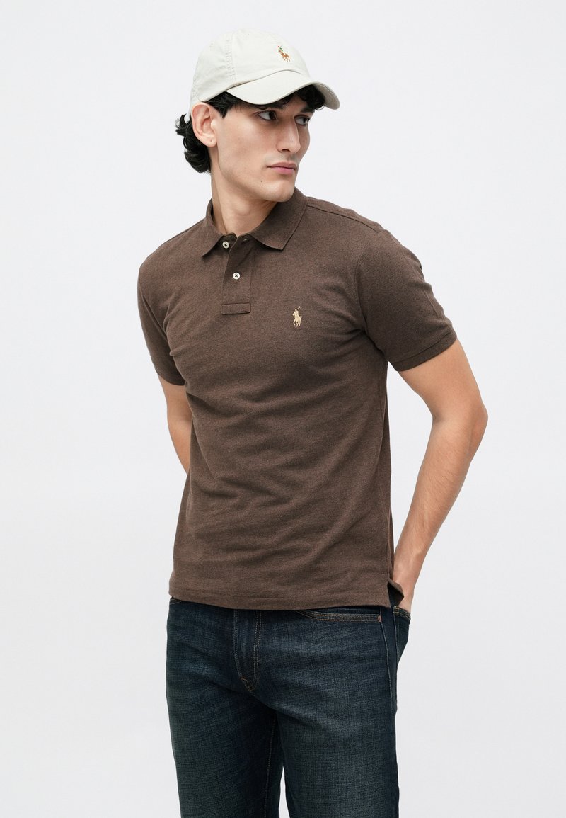 Ein junger Mann trägt ein braunes Poloshirt mit einem Logo, dunkle Jeans und eine beige Mütze. Er schaut zur Seite vor einem schlichten Hintergrund.