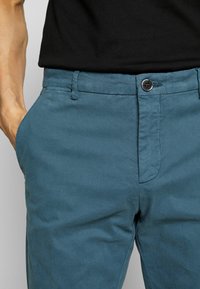 Chino broeken in teal, gemaakt van katoen. Kenmerken zijn een zachte textuur, voorzakken, riemlussen en een enkele knoopsluiting.