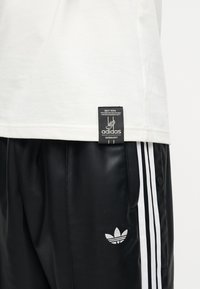 Fehér ing fekete Adidas Germany címkével, fekete nadrág fehér Adidas csíkokkal és Trefoil logóval a bal lábon.