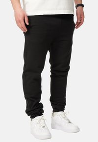 Black sweatpants feature a relaxed fit, ribbed ankle cuffs, and a smooth texture. Paired with white sneakers for contrast.
