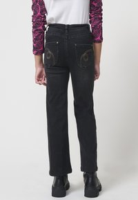 Los jeans negros tienen un corte recto, una cintura alta y costuras decorativas en color dorado en los bolsillos traseros. Se llevan con una camiseta blanca y botas negras.