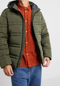 Oliwkowa puchowa kurtka z kapturem, pikowany fason, zapinana na zamek z przodu, noszona na rustykalnej koszuli na guziki. Widać niebieskie dżinsy.