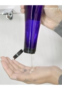 Lila Plastikflasche mit schwarzem Deckel, die eine klare, zähe Flüssigkeit in eine Hand gießt, vor einem hellen Hintergrund.