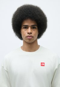 Light beige sweatshirt with round neck, featuring a small red logo. Model has a dense, round afro hairstyle. Plain background.