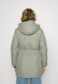 Giacca parka in verde oliva con cappuccio, vita svasata e una texture liscia, leggermente lucida. Presenta polsini elastici e un orlo posteriore più lungo.