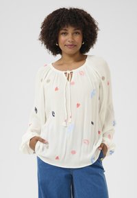 White, long-sleeved blouse with a round neckline and tie detail. Features colorful embroidered shapes, including hearts and flowers. Textured fabric.