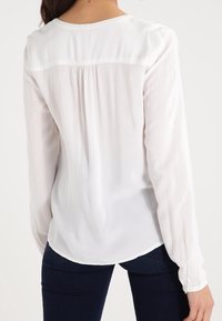 Vit långärmad blus med en len, lätt textur. Har en något rynkad bakdel och rundad nederkant. Kombineras med mörkblå jeans.