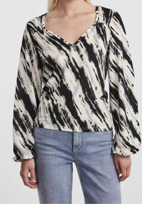 Långärmad topp i svart och krämfärgad vågmönster, med V-ringning och lätt insydda axlar, kombinerad med ljusblå jeans.