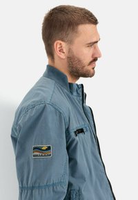 Blauwe bomberjack met een ritssluiting aan de voorkant, geribde kraag en een geborduurde patch met een landschapsontwerp op de linker mouw.