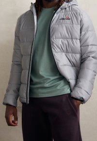 Helle graue Daunenjacke mit Kapuze und Reißverschluss, mit horizontalen Nähten, getragen über einem strukturierten grünen Oberteil und dunkelvioletten Hosen.