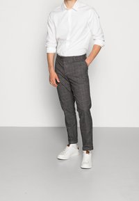 Hombre vestido con una camisa blanca de botones y las mangas remangadas, pantalones de cuadros grises y zapatillas blancas, de pie frente a un fondo sencillo.