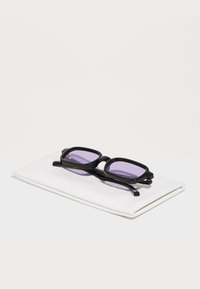 Gafas de sol negras con lentes de tintado púrpura, de forma rectangular y con montura gruesa, reposando sobre una funda blanca y suave.
