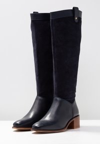 Knäboots med skaft i marinblå mocka och en slät lädertå. Har en låg träklack och ett dekorativt läderrem upptill.