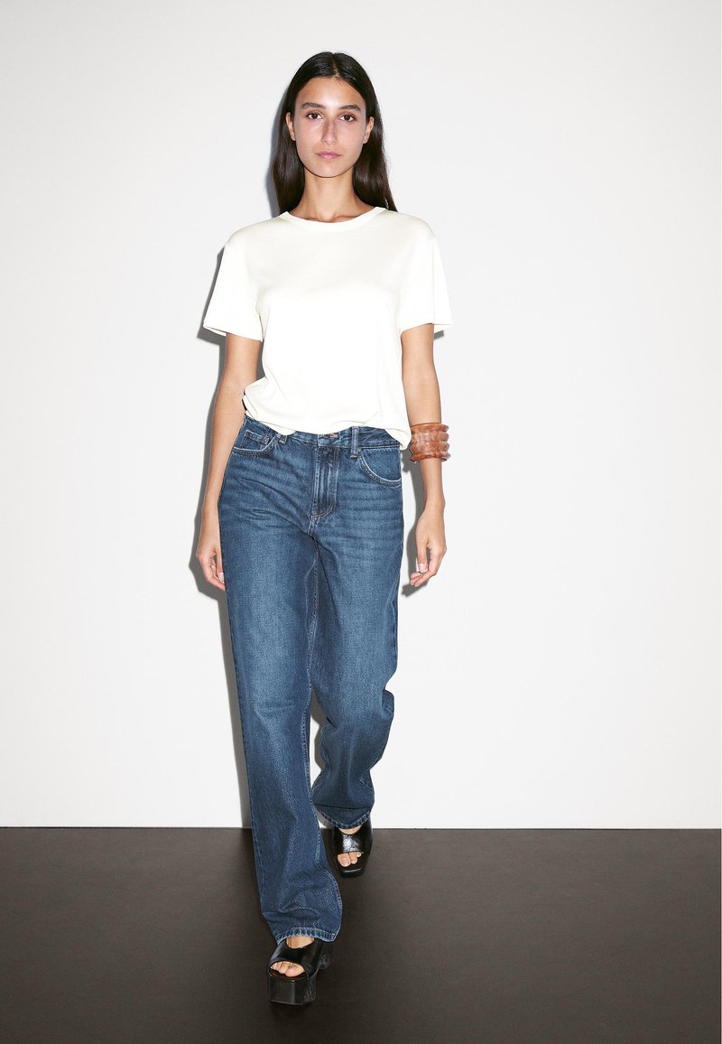 T-shirt bianco maniche corte, jeans in denim blu a gamba larga, braccialetto marrone al polso e scarpe con platforma nere. Outfit semplice e casual.