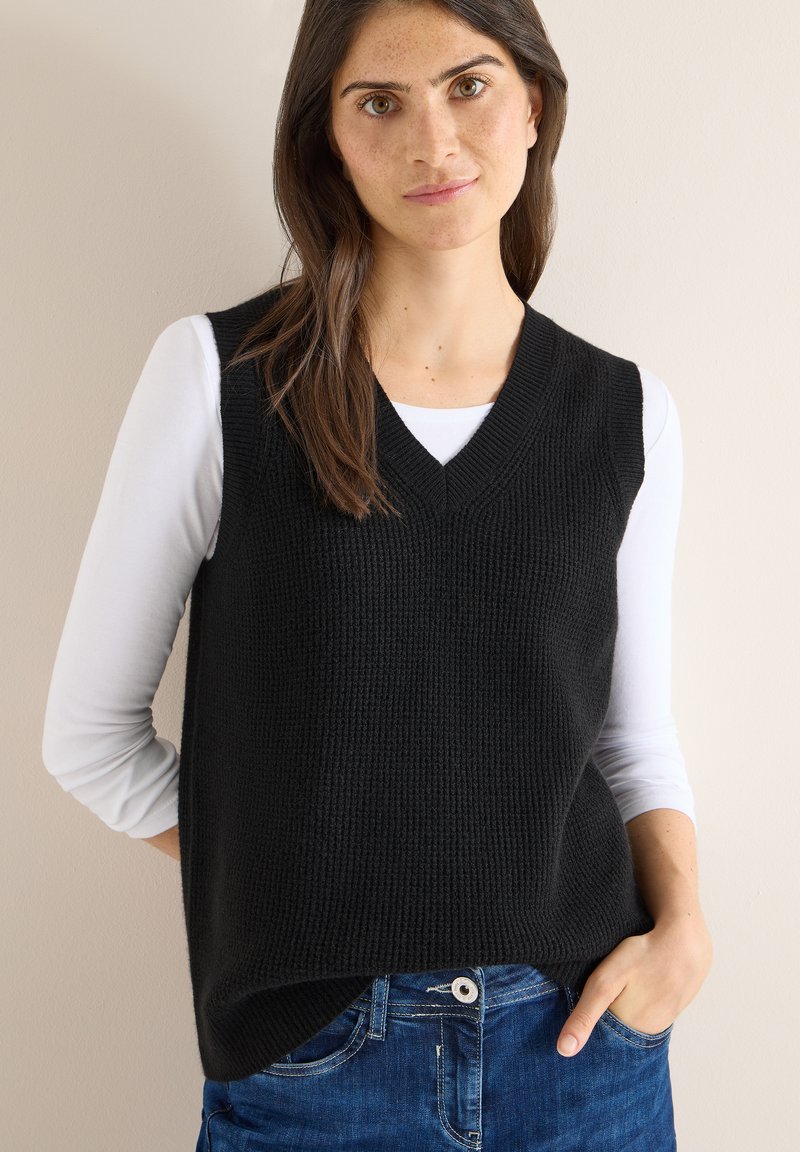 Schwarzer, gestrickter, ärmelloser Pullover mit V-Ausschnitt, strukturierter Stoff, getragen über einem weißen Langarmhemd. Kombiniert mit blauen Jeans.