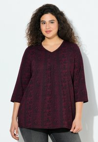 Haut bordeaux avec un col en V, manches trois-quarts, présentant un motif texturé et des détails de plis, associé à un jeans gris foncé.