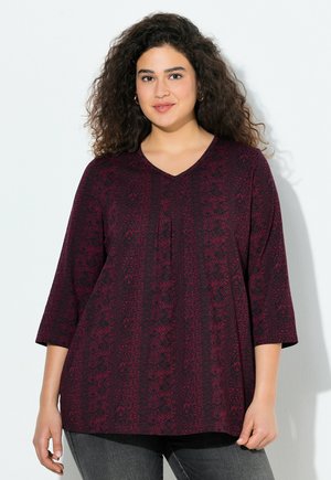 Haut bordeaux avec un col en V, manches trois-quarts, présentant un motif texturé et des détails de plis, associé à un jeans gris foncé.
