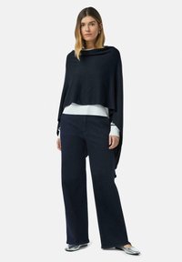 Kobieta ubrana w granatowy poncho, białą bluzkę z długim rękawem, ciemne szerokie spodnie i srebrne płaskie buty stoi na tle białym.