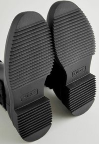 Suelas de botas de goma negra con textura estriada y forma ovalada. Presenta un logotipo "NEXT" y un tacón plano con un patrón claro para tracción.