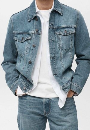 Personne portant une veste en jean bleue et un jean avec une chemise blanche à boutons et un t-shirt blanc, mains dans les poches de la veste.