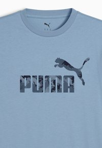 Šviesiai mėlyna marškinėliai su tamsiai mėlynu Puma logotipu, vaizduojančiu šuoliuojantį pumą, ir ryškiais blokiniais rašmenimis, užrašytais "PUMA" per krūtinę.