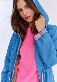 Niebieska kurtka z kapturem i sznurkiem, z różowymi i szarymi akcentami, na różowym swetrze. Gładki materiał z luźnym krojem i zapięciem na zamek.