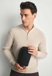 Beżowy sweter z prążkowanej tkaniny z kołnierzem na połowę zamka; osoba trzyma czarną torbę o fakturze z wyraźnym zamkiem błyskawicznym i subtelnym detalem logo.