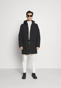 Svart parkajakke med hette, frontlukking med knapper, to sidelommer, brukt over en lys turtleneck, kombinert med hvite bukser og svarte støvler.