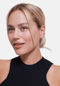 Zwarte geribbelde hoogsluitende top, model met blond haar en lichte huid, draagt kleine gouden ringoorbellen met een subtiele textuur en glans.
