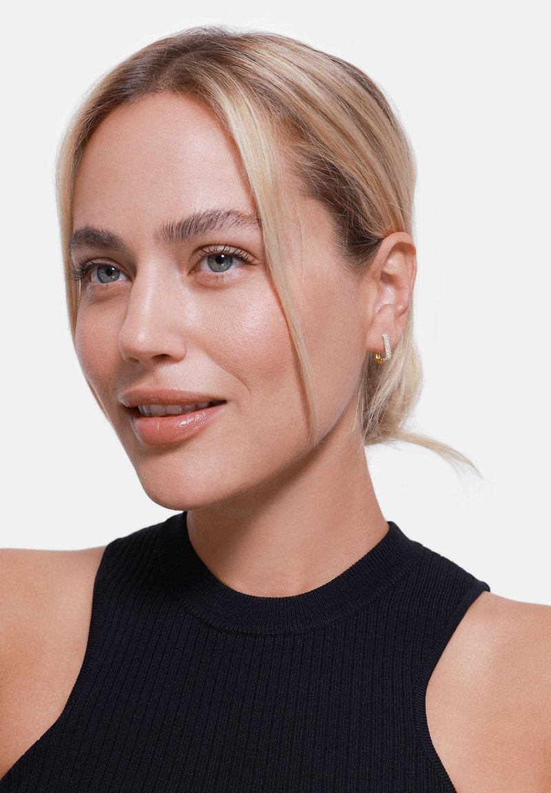 Zwarte geribbelde hoogsluitende top, model met blond haar en lichte huid, draagt kleine gouden ringoorbellen met een subtiele textuur en glans.