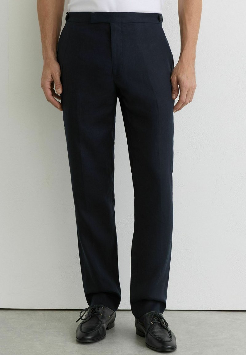Homme portant un pantalon noir sur mesure, des chaussures noires à lacets, et une chemise blanche, debout contre un mur clair et un sol gris.