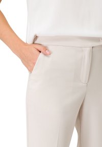 Pantalon beige avec une texture lisse, doté d'une poche latérale et d'une taille ajustée, associé à un haut blanc.