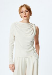 Haut crème drapé avec une longue manche, présentant un petit accent doré sur l'épaule. Tissu doux avec une texture lisse.