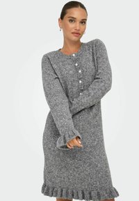 Femme portant une robe en tricot gris à manches longues avec des boutons sur le devant et des poignets et un ourlet à volants, se tenant devant un fond blanc.