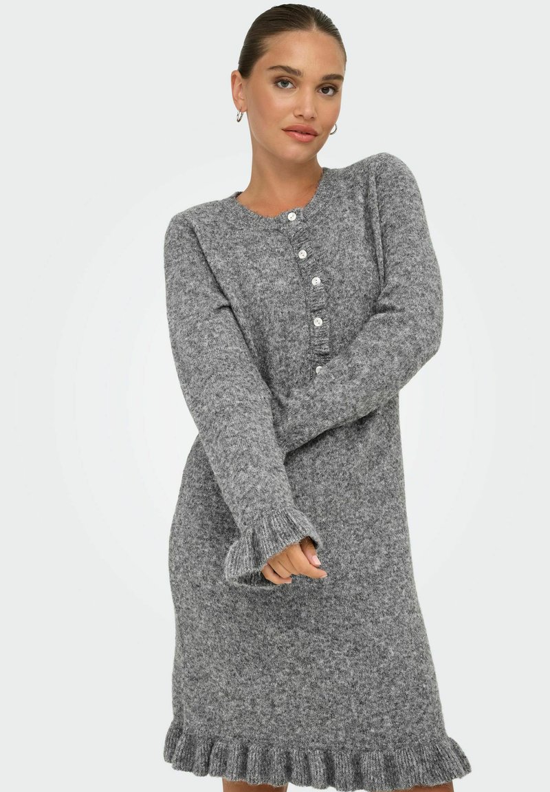 Femme portant une robe en tricot gris à manches longues avec des boutons sur le devant et des poignets et un ourlet à volants, se tenant devant un fond blanc.