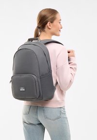 Grå ryggsäck med en slät yta, som har en framficka, justerbara remmar och ett robust handtag upptill, bärs över en rosa tröja.