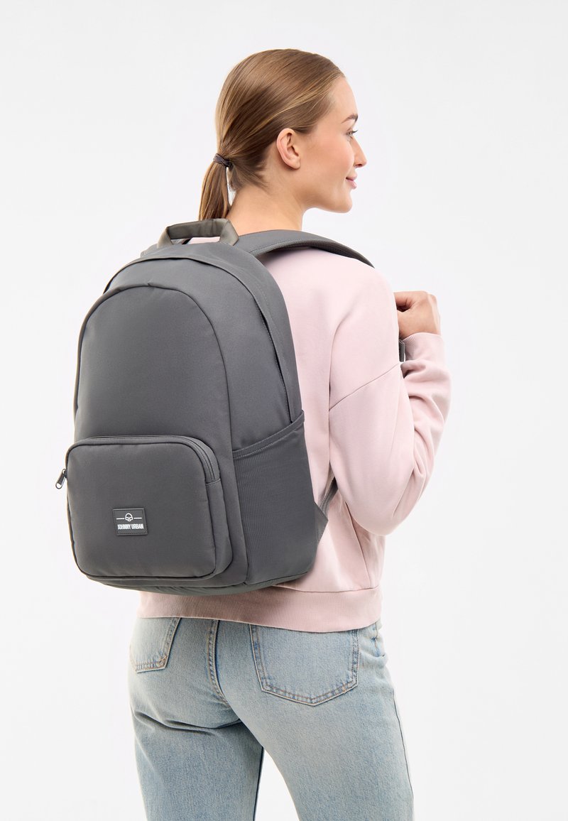 Grå ryggsäck med en slät yta, som har en framficka, justerbara remmar och ett robust handtag upptill, bärs över en rosa tröja.