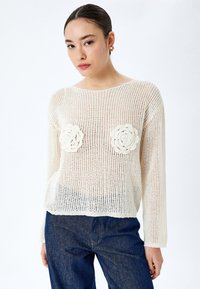 Luftiger cremefarbener Pullover mit floralen Häkelakzenten auf der Brust. Hat lange Ärmel und einen weiten Ausschnitt, kombiniert mit dunkelblauen Jeans.